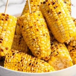 Bowl filled with grilled corn on the cob halves on sticks, ready to serve with salt and pepper in the background.