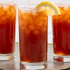 Three tall glasses of refreshing iced tea with ice cubes and lemon wedges on the rim are arranged on a metal tray, surrounded by lemon slices and a bowl of whole lemons—a perfect showcase for your favorite iced tea recipe.