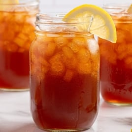 Three mason jars filled with Iced Tea and ice cubes, each garnished with a slice of lemon on the rim, are placed on a white surface with lemon wedges nearby&mdash;perfect for trying out your favorite iced tea recipe.