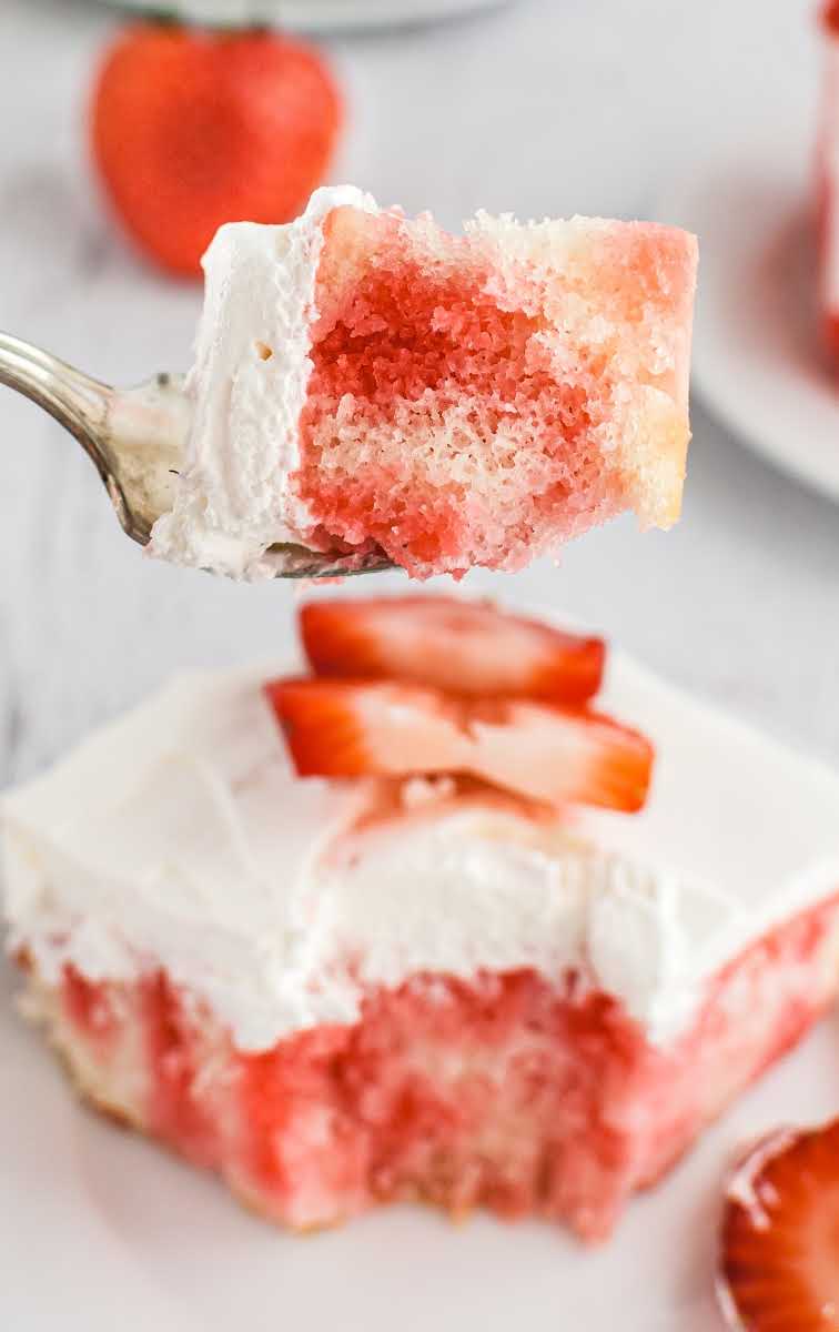 close up shot of a slice of jello poke cake topped with whipped cream and strawberry slices with a piece on a fork