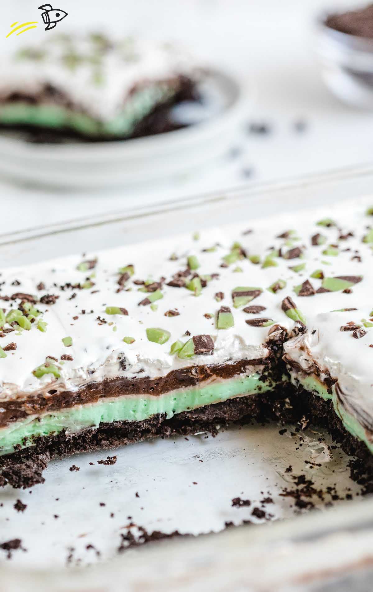 Partially sliced mint chip dessert lasagna in a glass dish showing colorful layers and crumbled Oreo base.\