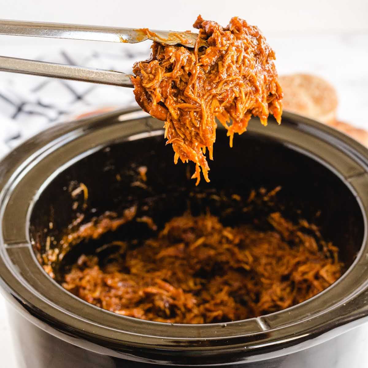 close up shot of pulled bbq chicken in a slow cooker with a piece picked up with tongs