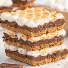 Stack of gooey s’mores bars with layers of toasted marshmallows, melted chocolate, and graham cracker crust on a white surface.
