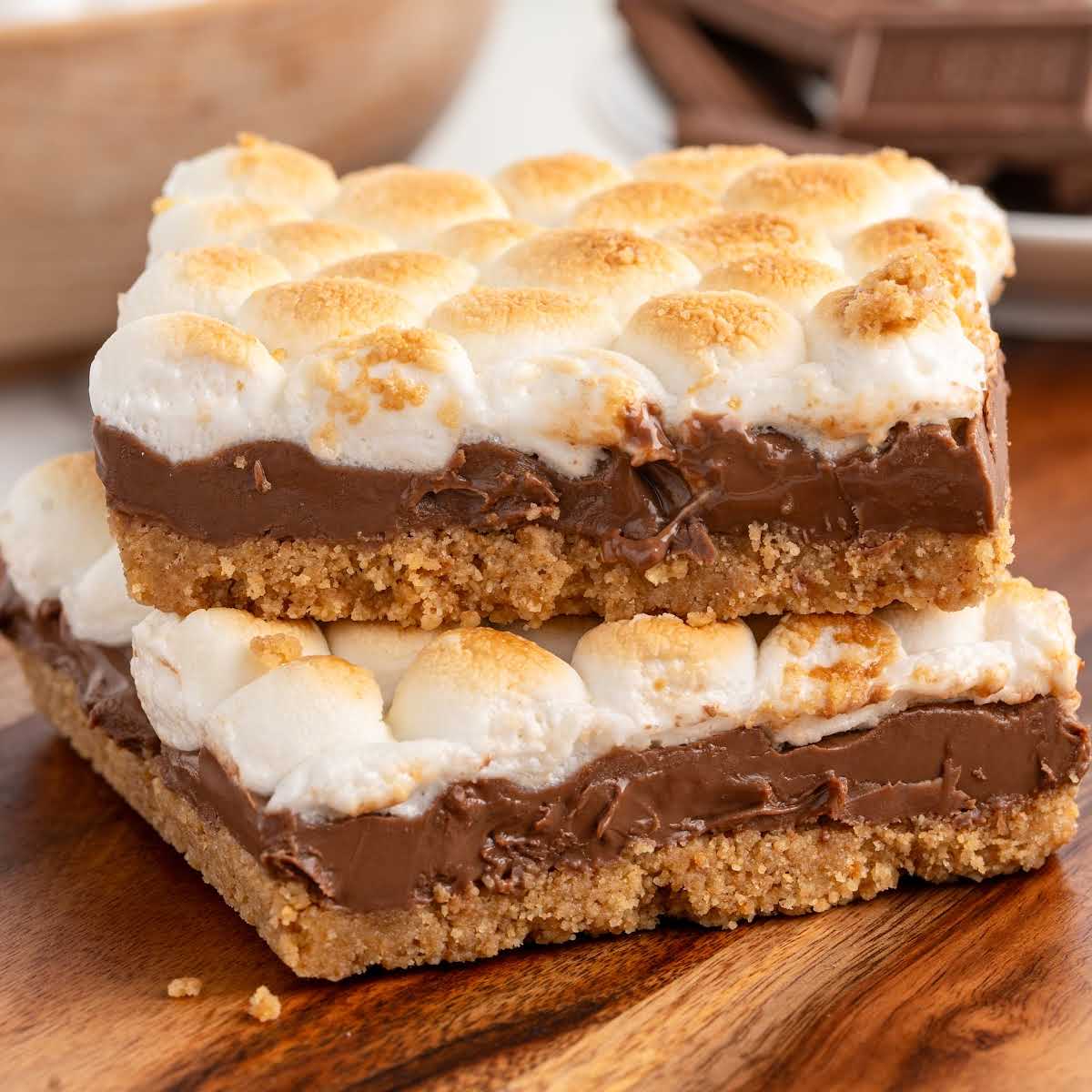 Two s’mores bars stacked on a wooden board with gooey chocolate and toasted marshmallows on a graham cracker crust.