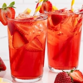 Two tall glasses of iced strawberry sweet tea surrounded by fresh strawberries and lemon wedges.