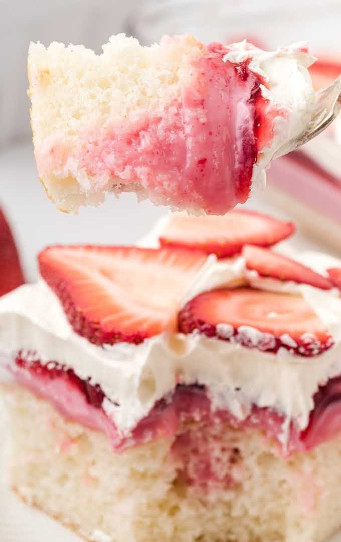close up shot of a slice of strawberry poke cake topped with whipped cream and strawberry slices with a piece being taken out of it with a fork
