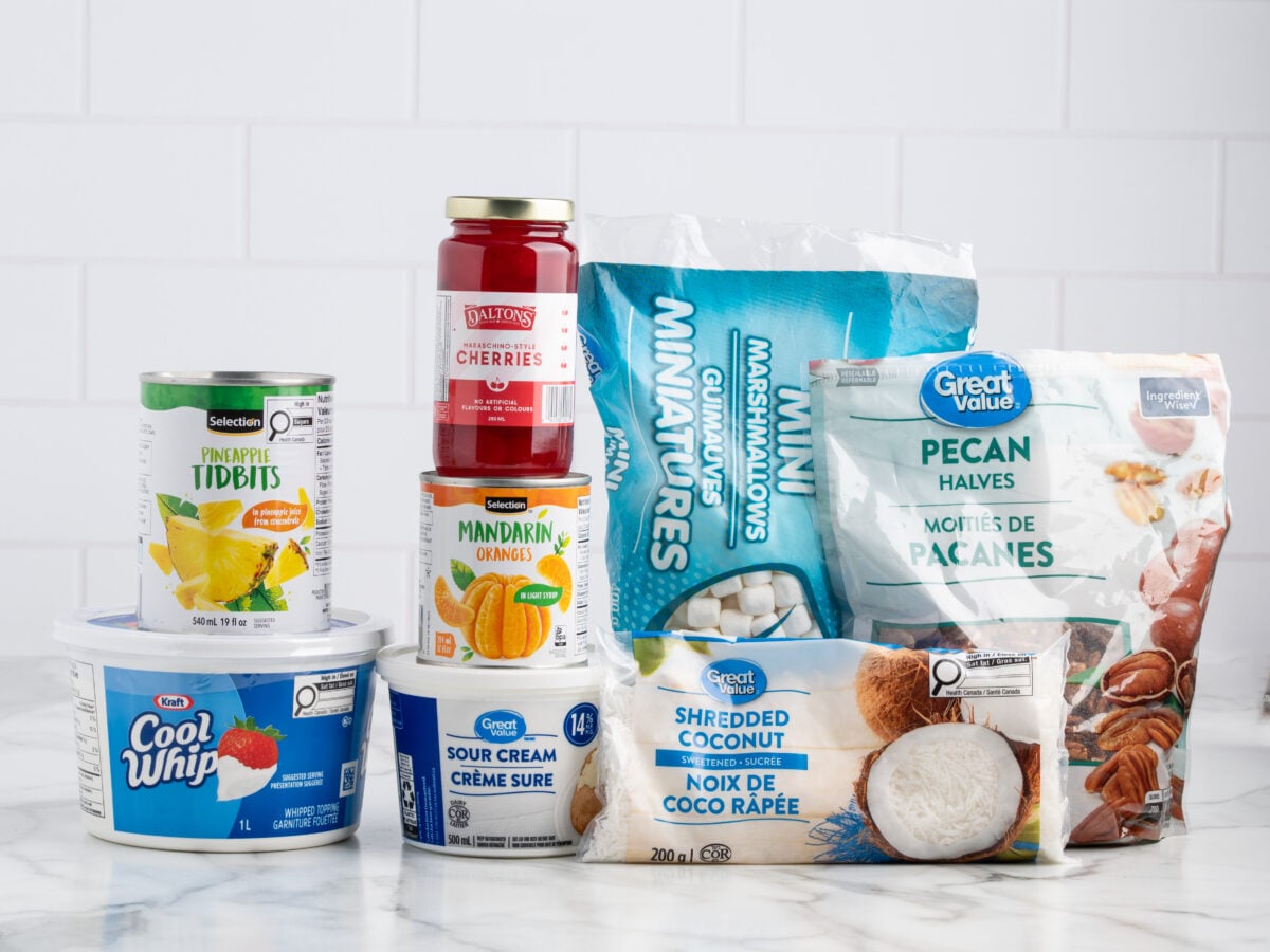 A selection of Ambrosia Salad ingredients on a marble counter, including Cool Whip, sour cream, pineapple tidbits, mandarin oranges, cherries, mini marshmallows, pecan halves, and shredded coconut, with a white tiled background.
