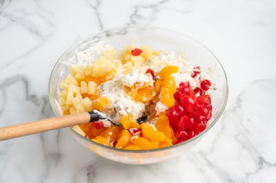 A glass bowl on a marble surface contains Ambrosia Salad ingredients: whipped cream, chopped pineapples, mandarin orange slices, and maraschino cherries, with a wooden spoon ready for mixing.
