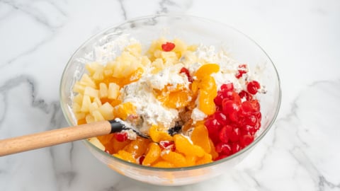 A glass bowl on a marble surface contains Ambrosia Salad ingredients: whipped cream, chopped pineapples, mandarin orange slices, and maraschino cherries, with a wooden spoon ready for mixing.
