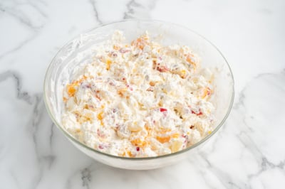 A glass bowl filled with a creamy Ambrosia Salad mixture, containing pieces of orange and red fruit, set on a white marble surface.