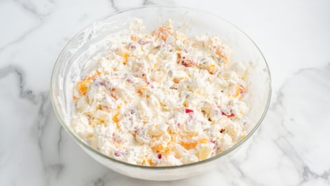 A glass bowl filled with a creamy Ambrosia Salad mixture, containing pieces of orange and red fruit, set on a white marble surface.