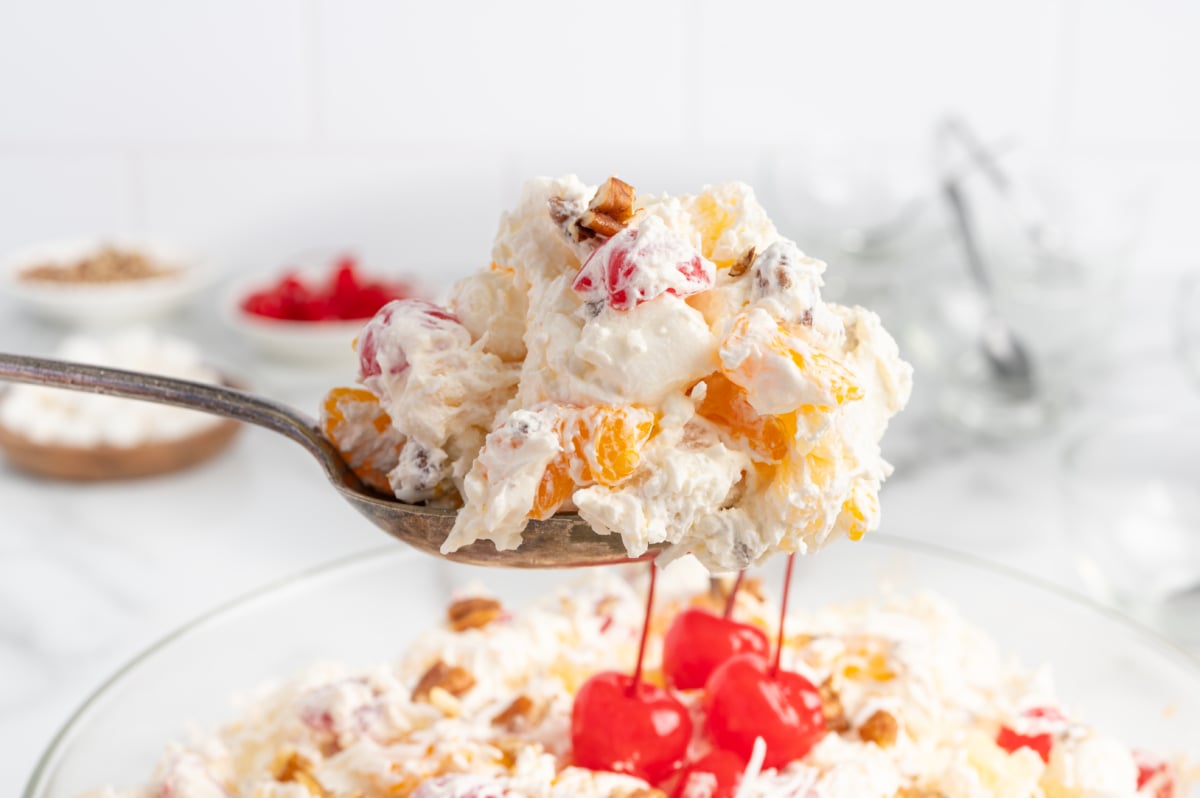 A close-up of a spoonful of Ambrosia Salad with whipped cream, mandarin oranges, pineapple, cherries, coconut, and nuts, held above a bowl filled with the classic dessert garnished with maraschino cherries.
