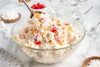 A large glass bowl brimming with Ambrosia Salad—whipped cream, marshmallows, cherries, pineapple, coconut, and nuts—invites you to scoop some out. Ingredients and empty glass dishes are arranged around the colorful dessert.