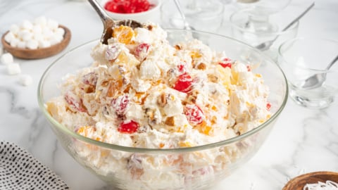 A large glass bowl brimming with Ambrosia Salad&mdash;whipped cream, marshmallows, cherries, pineapple, coconut, and nuts&mdash;invites you to scoop some out. Ingredients and empty glass dishes are arranged around the colorful dessert.