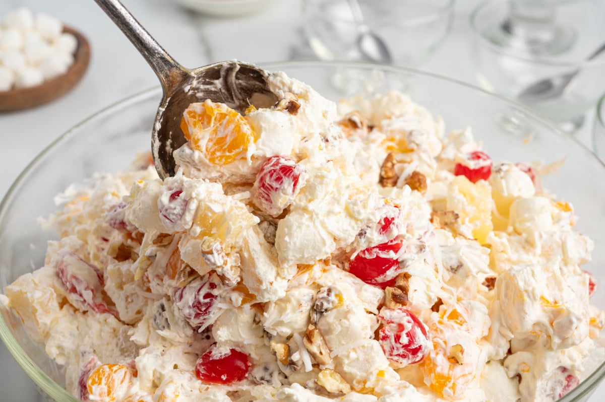 A large glass bowl filled with creamy Ambrosia Salad, featuring whipped cream, cherries, pineapple, orange segments, and chopped nuts, with a serving spoon scooping some out.