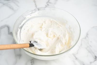 A glass bowl filled with fluffy whipped cream for Ambrosia Salad, with a spatula resting inside, sits on a white marble surface.