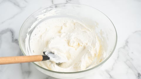 A glass bowl filled with fluffy whipped cream for Ambrosia Salad, with a spatula resting inside, sits on a white marble surface.