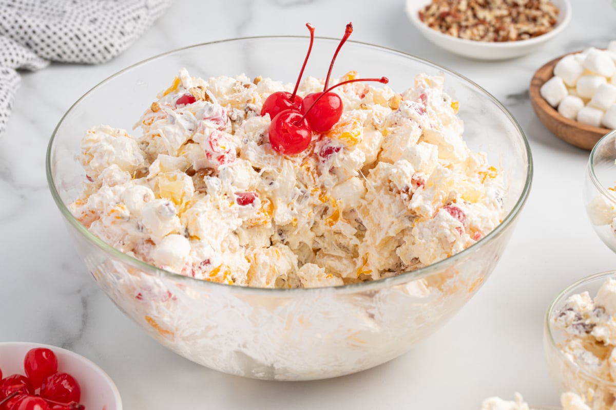 A glass bowl filled with creamy Ambrosia Salad topped with three maraschino cherries, surrounded by bowls of mini marshmallows, pecans, and cherries on a white marble surface.