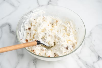 A glass bowl filled with creamy Ambrosia Salad, dotted with small fruit chunks and being mixed with a wooden spoon, sits on a white marble surface.