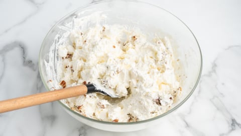 A glass bowl filled with creamy Ambrosia Salad, dotted with small fruit chunks and being mixed with a wooden spoon, sits on a white marble surface.