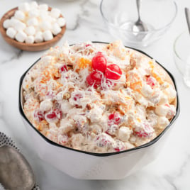 A large white bowl of creamy Ambrosia Salad topped with maraschino cherries and shredded coconut, surrounded by bowls of mini marshmallows, pecans, cherries, and coconut on a marble surface.
