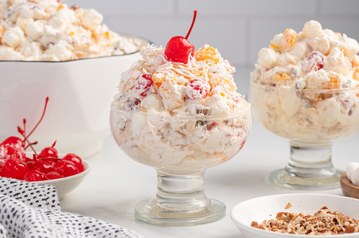 Two glass dessert bowls filled with creamy Ambrosia Salad, topped with shredded coconut and a maraschino cherry, sit in front of a larger bowl of Ambrosia Salad garnished with extra cherries and pecans in the background.