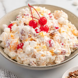 A bowl of Ambrosia Salad topped with maraschino cherries, featuring whipped cream, marshmallows, orange segments, pineapple, and chopped nuts on a light surface.