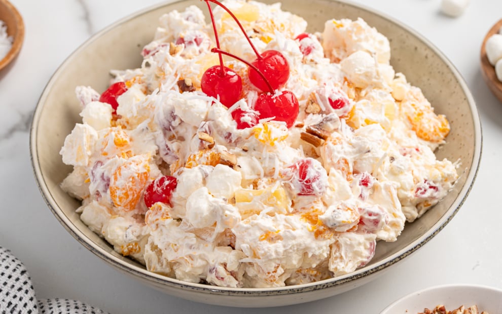 A bowl of Ambrosia Salad topped with maraschino cherries, featuring whipped cream, marshmallows, orange segments, pineapple, and chopped nuts on a light surface.