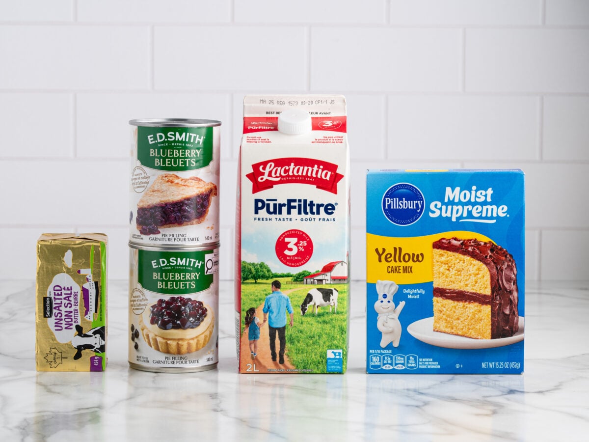 Five baking ingredients on a counter&mdash;perfect for Blueberry Dump Cake: butter, two cans of blueberry pie filling, a carton of 3% milk, and Pillsbury Moist Supreme yellow cake mix&mdash;all set against a white tiled background.