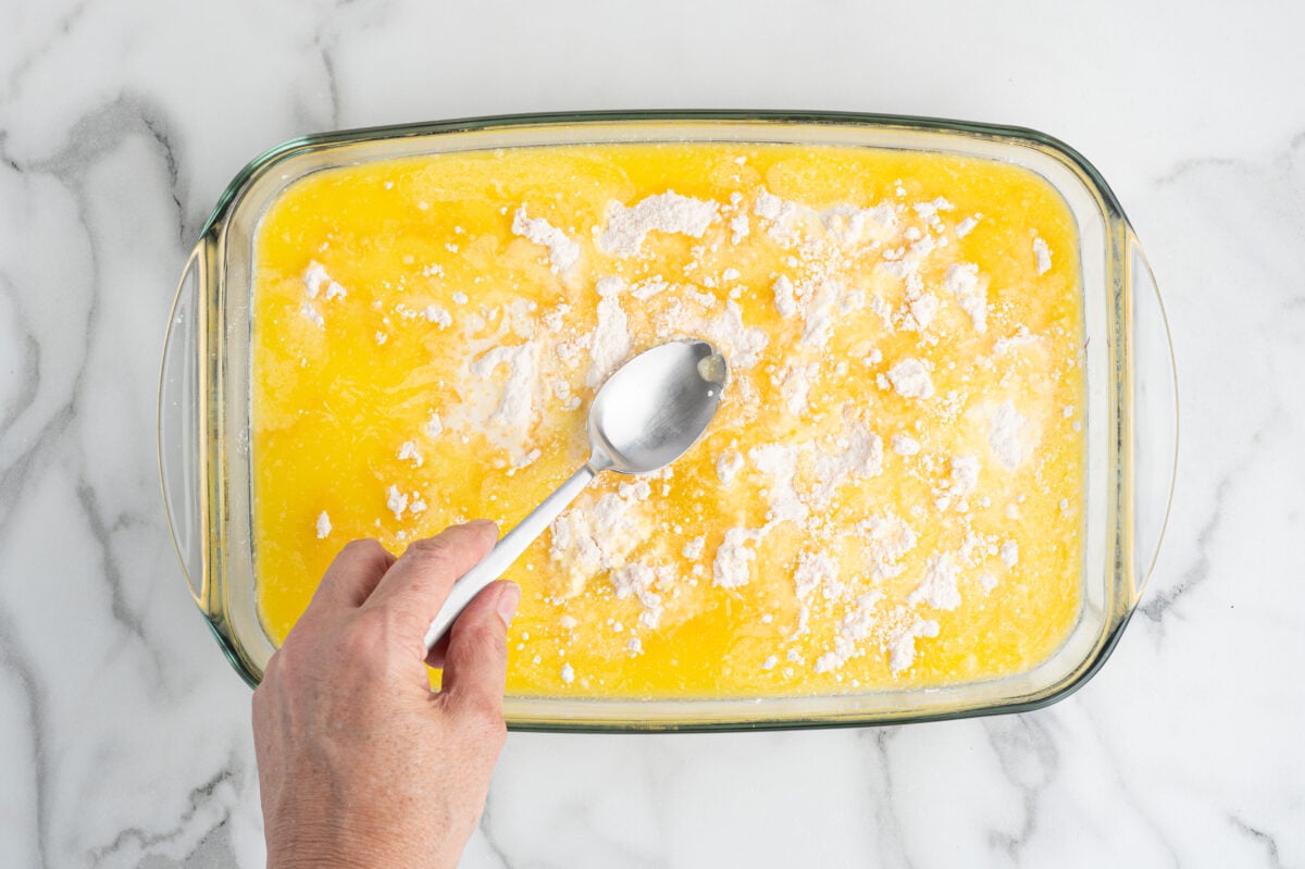A hand holding a spoon is stirring a glass baking dish filled with a yellow liquid mixture and sprinkled white flour on top, preparing a delicious Blueberry Dump Cake on a marble surface.