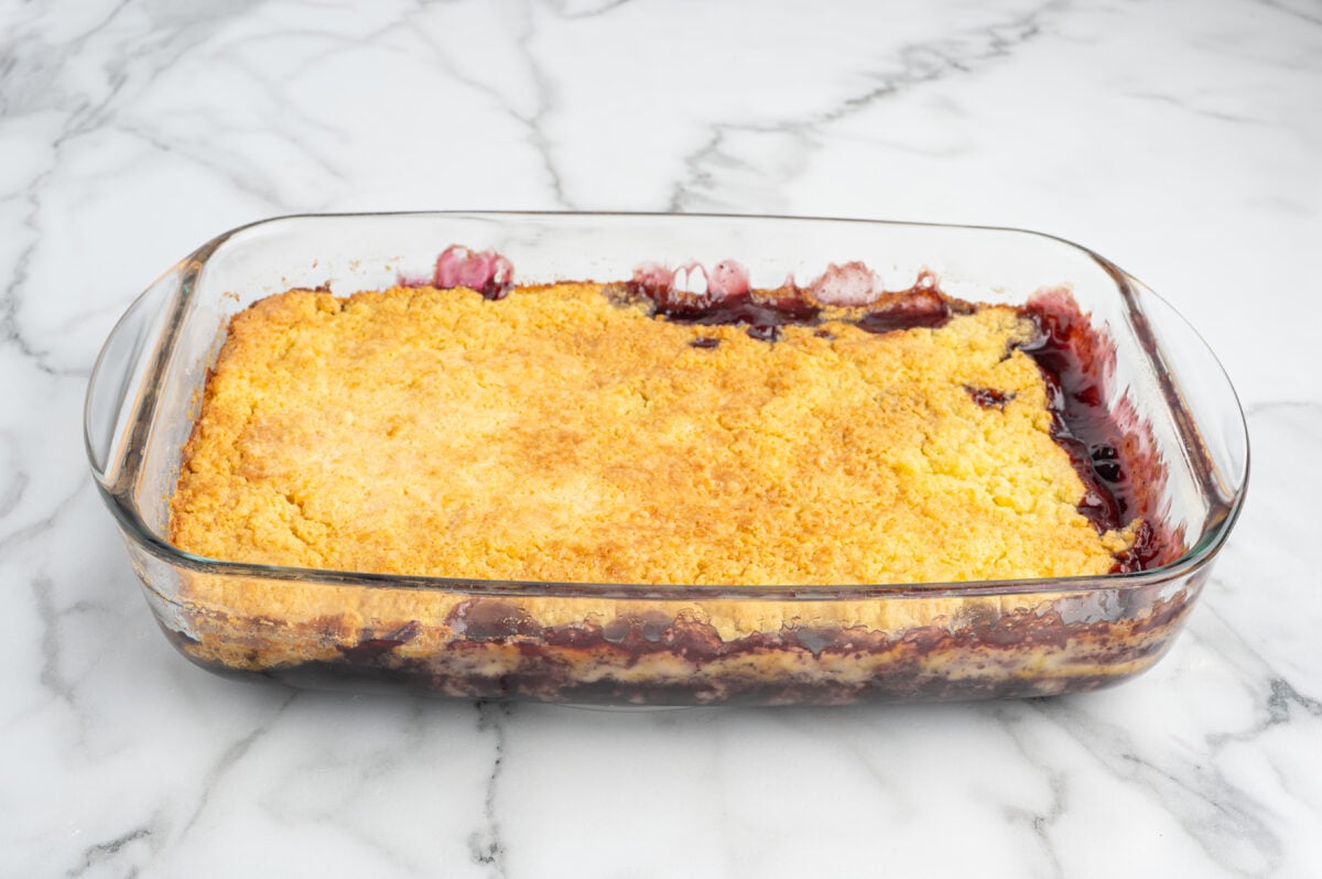 A glass baking dish filled with Blueberry Dump Cake showcases a golden brown crust on top and bubbling, dark fruit filling peeking around the edges. The dish is set on a white marble surface.