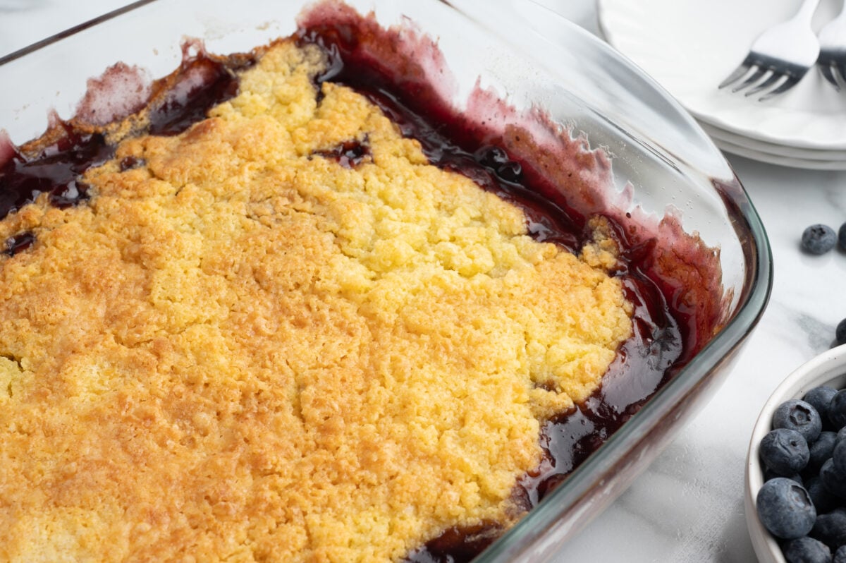 A close-up of a freshly baked Blueberry Dump Cake in a glass baking dish, with a golden, crumbly topping and bubbling blueberry filling around the edges. Nearby are plates, forks, and a bowl of fresh blueberries.