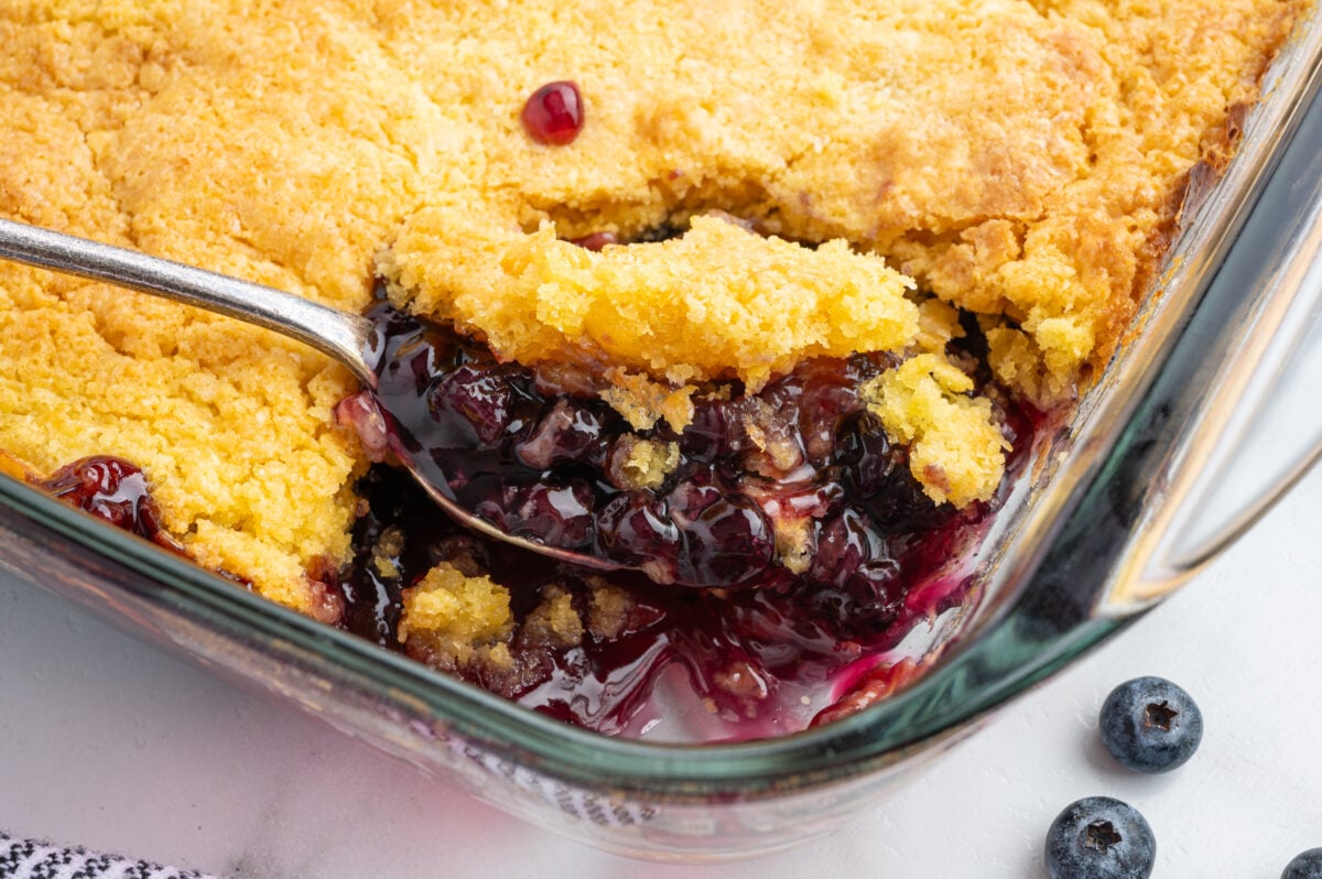 A close-up of a glass baking dish filled with blueberry cobbler, also known as Blueberry Dump Cake. A spoon scoops out a portion, revealing juicy blueberries and thick purple filling beneath the golden, crumbly crust. Fresh blueberries are nearby.