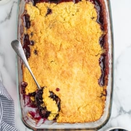 A glass baking dish filled with golden-brown Blueberry Dump Cake sits on a white surface. A serving spoon rests in one corner, revealing dark, juicy berries beneath the crisp topping.