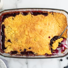 A glass baking dish filled with golden-brown Blueberry Dump Cake, with a spoon scooping out a portion. Fresh blueberries, glasses of milk, plates, and utensils are arranged nearby on a marble surface.