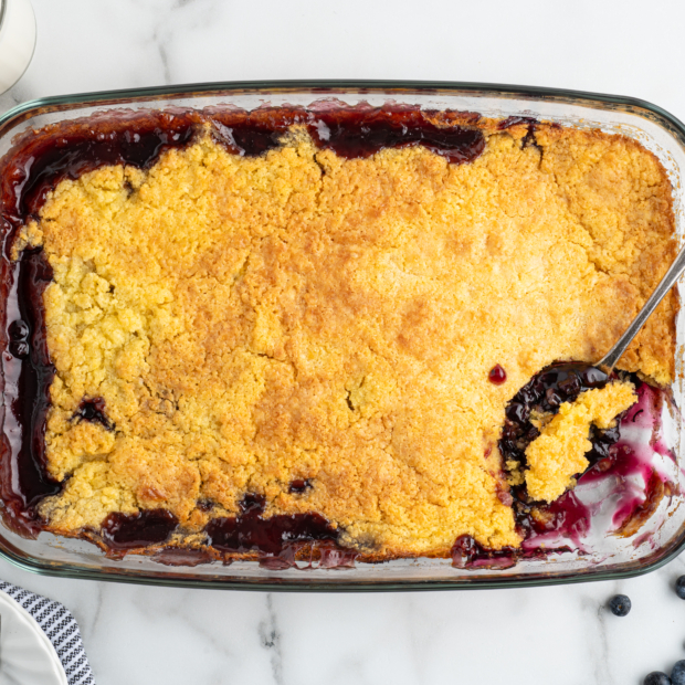 A glass baking dish filled with golden-brown Blueberry Dump Cake, with a spoon scooping out a portion. Fresh blueberries, glasses of milk, plates, and utensils are arranged nearby on a marble surface.
