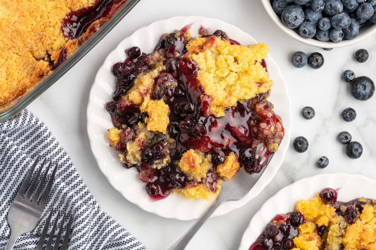 A plate with a serving of blueberry cobbler, similar to Blueberry Dump Cake, topped with a golden crumbly crust, surrounded by fresh blueberries, a baking dish, and a striped napkin with forks on a white surface.