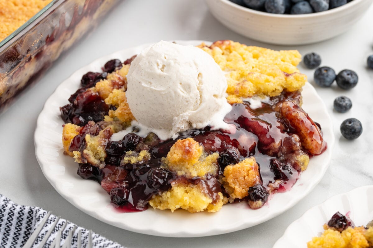 A plate of blueberry cobbler, reminiscent of Blueberry Dump Cake, is topped with a scoop of vanilla ice cream on a white table. Fresh blueberries and a baking dish filled with more cobbler are in the background.