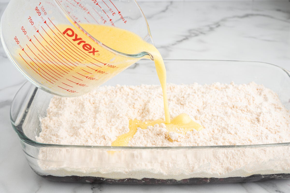 A glass measuring cup pours a yellow liquid mixture onto dry flour in a rectangular glass baking dish on a marble countertop, perfect for making Blueberry Dump Cake.
