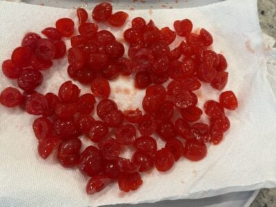 Bright red maraschino cherries, halved and pitted, are spread out on a white paper towel to dry&mdash;perfectly glossy and vibrant for adding to Ambrosia Salad, with the towel showing a few cheerful red juice stains.