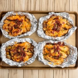Four Chicken Foil Packets filled with diced potatoes, chicken, melted cheese, bacon bits, and chopped chives sit on a baking sheet atop a wooden surfaceโan easy chicken foil packets recipe for any night.