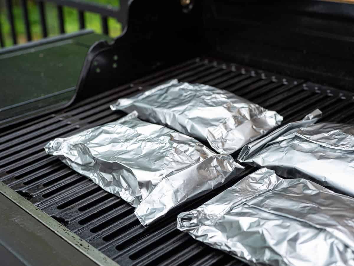 Four Chicken Foil Packets are placed on the grates of an outdoor grill, ready to be cooked. The grill lid is open, and the background shows greenery—a perfect setting for this easy chicken foil packets recipe.