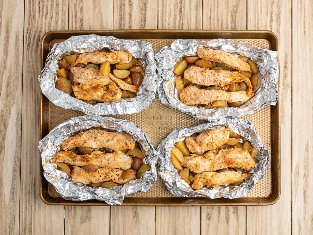 Four Chicken Foil Packets on a baking sheet, each filled with seasoned chicken strips and diced potatoes, are baked to perfection and arranged on a wooden surface—a delicious chicken foil packets recipe for an easy meal.