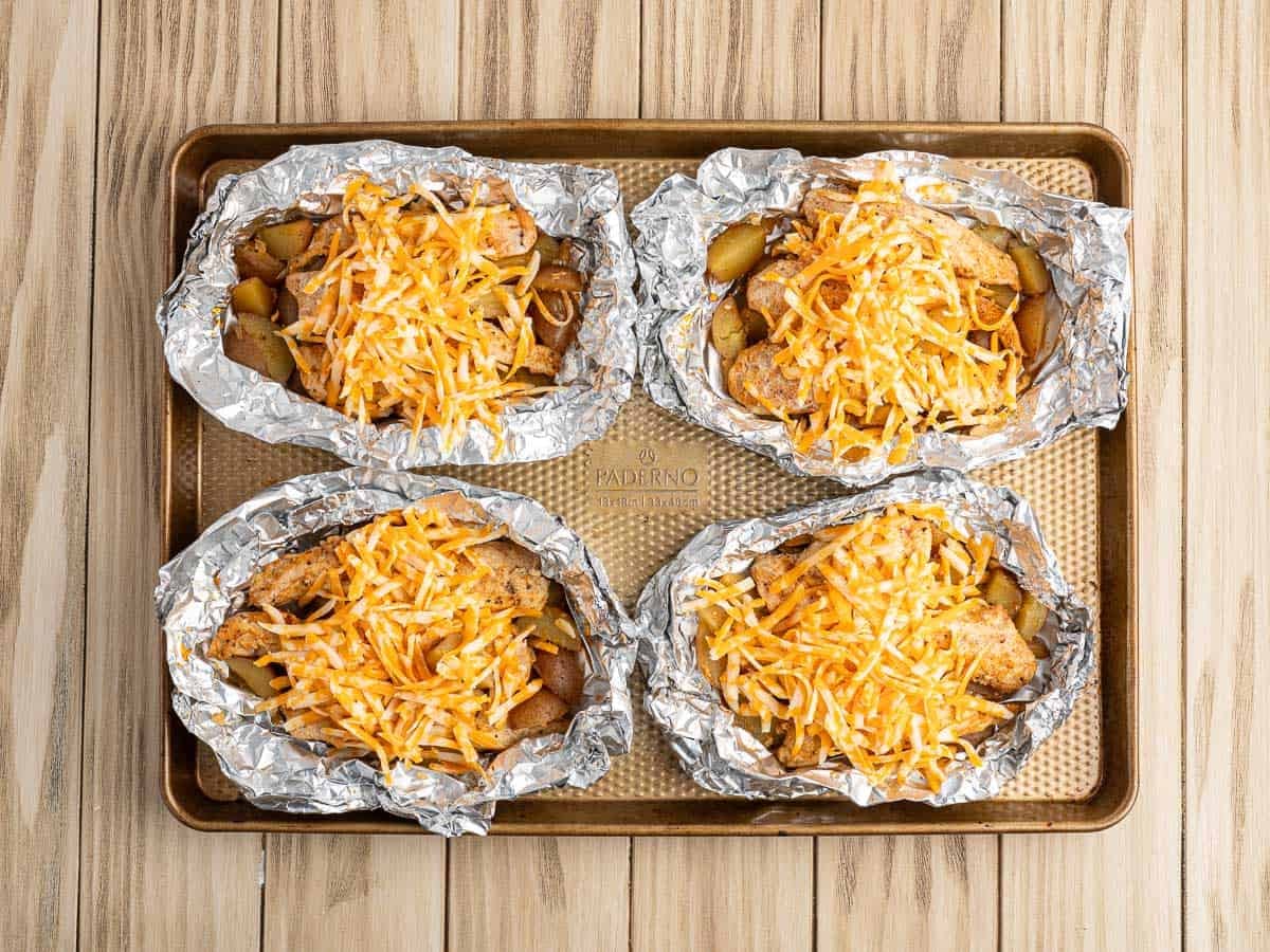 Four chicken foil packets filled with sliced potatoes and topped with shredded cheese sit on a baking sheet on a wooden surface, ready to be baked—a delicious chicken foil packets recipe in the making.