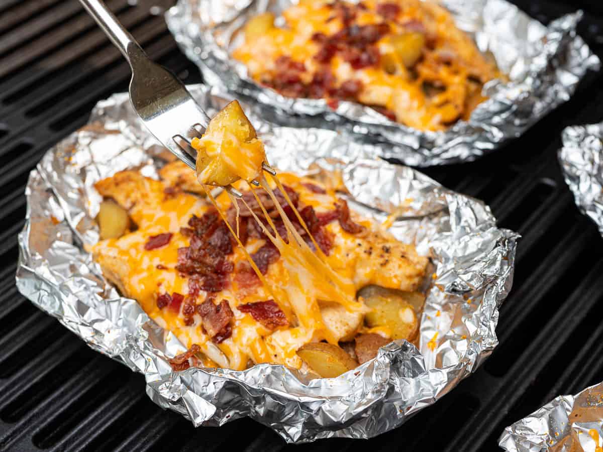 A fork lifts a cheesy, bacon-topped potato chunk from a foil packet, with melted cheese stretching, over more Chicken Foil Packets filled with potatoes, cheese, and bacon sizzling on the grill—perfect for any chicken foil packets recipe.