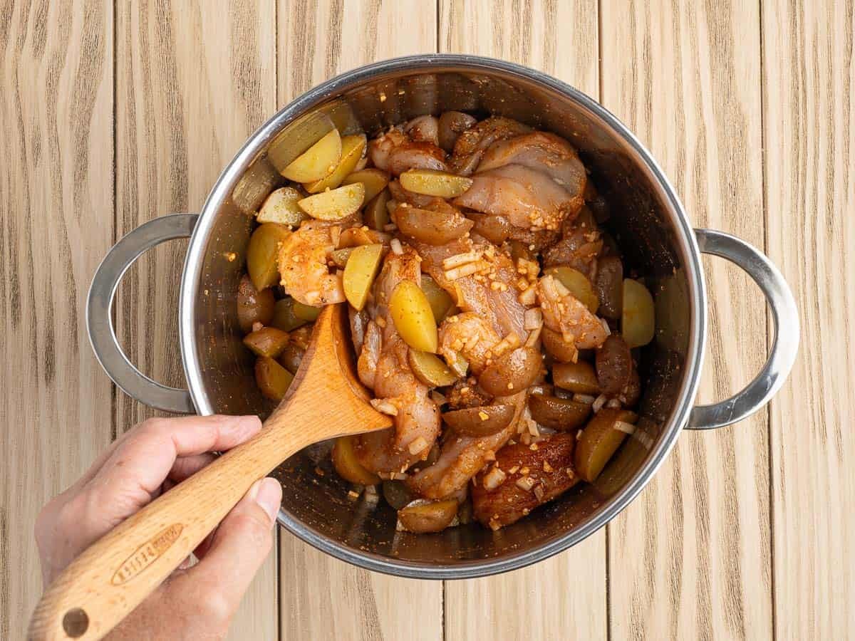 A hand stirs marinated raw chicken pieces, onions, and yellow vegetables in a large metal pot with a wooden spoon on a light wooden surface—perfect prep for a tasty Chicken Foil Packets recipe.