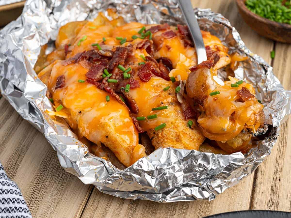 Baked Chicken Foil Packets recipe: chicken breast in foil topped with melted cheddar cheese, bacon bits, and chopped chives, with a fork taking a bite; wooden table background.