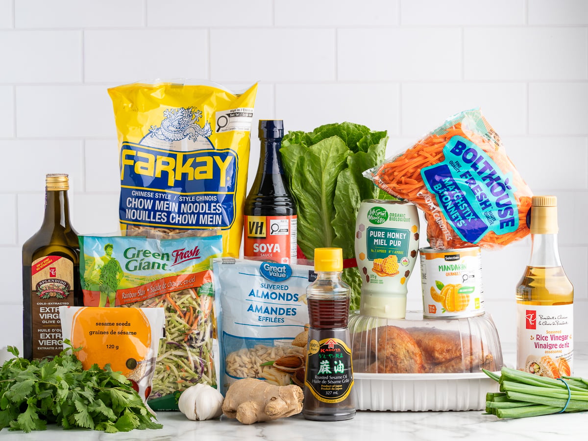 A variety of groceries on a counter, perfect for making Chinese Chicken Salad, including chow mein noodles, lettuce, rotisserie chicken, shredded carrots, soy sauce, olive oil, sesame oil, slivered almonds, ginger, honey, and green onions.
