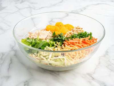 A glass bowl on a marble surface contains the vibrant ingredients of a Chinese Chicken Salad—shredded lettuce, chicken, mandarin oranges, green onions, carrots, cilantro, slivered almonds, and crunchy noodles—all arranged separately.