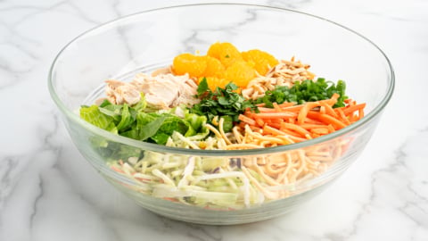 A glass bowl on a marble surface contains the vibrant ingredients of a Chinese Chicken Salad&mdash;shredded lettuce, chicken, mandarin oranges, green onions, carrots, cilantro, slivered almonds, and crunchy noodles&mdash;all arranged separately.